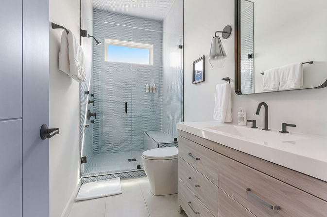 Bright modern coastal-style bathroom with glass-enclosed blue penny-tile shower and bench, small high window, light wood vanity with white countertop, matte-black faucets and hardware, wall mirror and white towels.