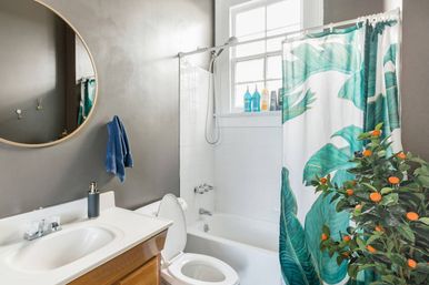Contemporary bathroom with white bathtub and tiled shower, teal tropical leaf shower curtain, potted plant with small orange fruits, round mirror, gray walls, window and blue hand towel