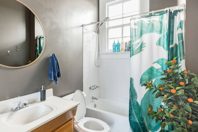 Contemporary bathroom with white bathtub and tiled shower, teal tropical leaf shower curtain, potted plant with small orange fruits, round mirror, gray walls, window and blue hand towel