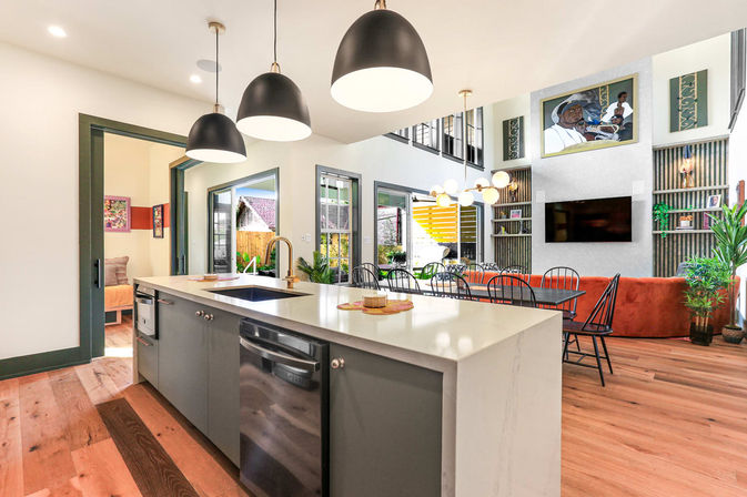 Bright modern open-concept kitchen and living room with white quartz island, brass faucet and built-in dishwasher, three black dome pendant lights, hardwood floors, dining table with black chairs, orange sofa, wall-mounted TV and tall windows overlooking a sunny backyard.