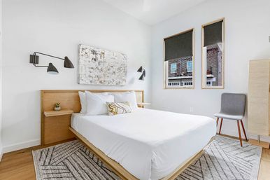 Bright, minimalist city apartment bedroom with white walls, wood platform bed and white linens, black swing-arm wall lamps, abstract wall art, two windows overlooking brick buildings, gray accent chair and patterned area rug