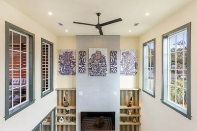 Sunlit double-height modern living room with tall green-trim windows, central gray feature wall topped by four colorful abstract geometric portraits, black ceiling fan, recessed TV and built-in wood shelving with decorative accents.