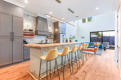 Open-plan modern kitchen and living area with a long wooden island and six yellow bar stools, gray cabinets, tiled backsplash, hardwood floors, high ceilings, teal sofa and sliding glass doors to a sunny patio.