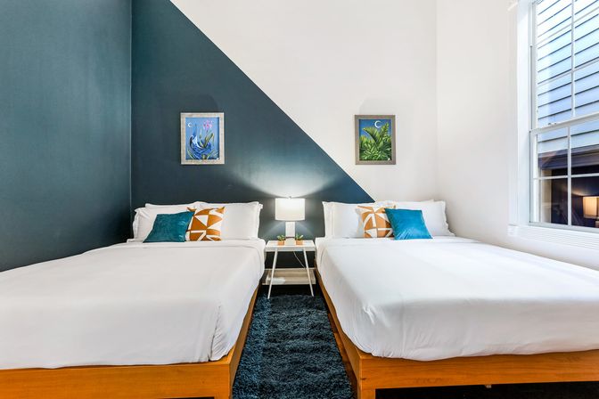 Bright modern twin bedroom with diagonal navy accent wall, two white beds with teal and orange pillows, a central nightstand with lamp, and a sunlit window