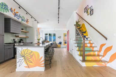 Open-concept modern kitchen and living area with long stainless island lined with black bar stools, gray cabinets, light wood floors, and a bright tropical mural wrapping the island and staircase for a playful coastal vibe.