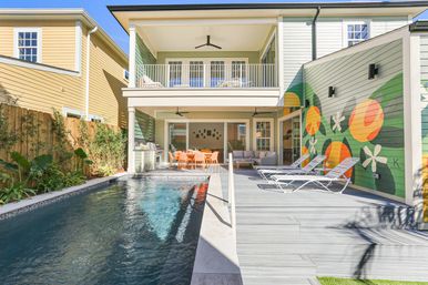 Sunny modern two-story backyard with narrow lap pool, gray wood deck, three white lounge chairs, bright orange dining set under a covered patio, upper balcony, and colorful fruit-and-flower mural on the exterior wall.