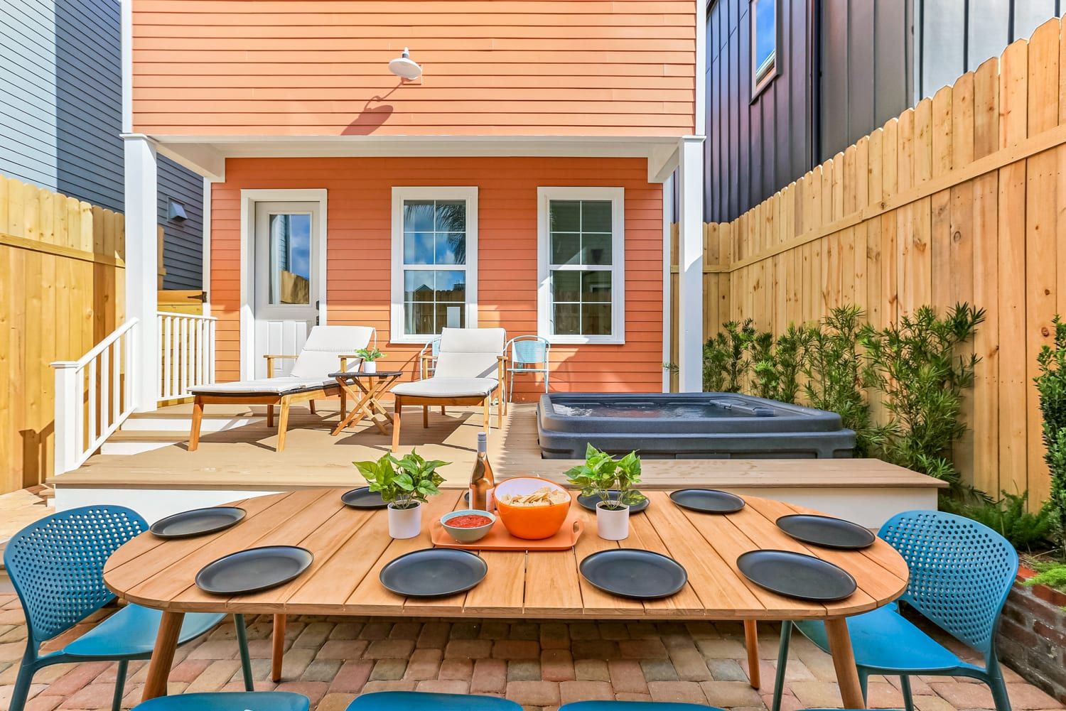 Cozy fenced backyard patio with wooden dining table and blue chairs, orange-sided house, lounge chairs and hot tub — ready for al fresco dining.