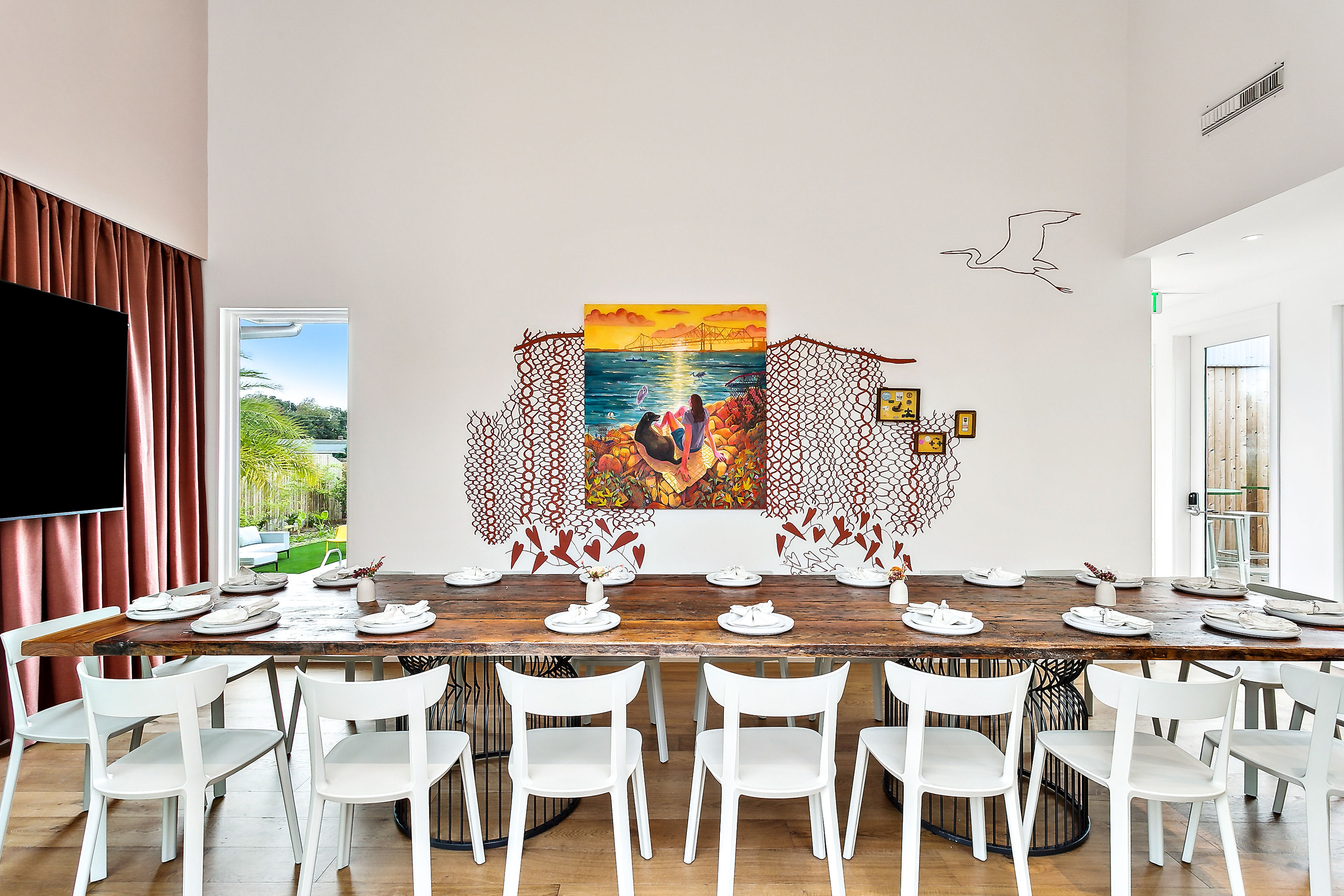 Bright modern coastal dining room with a long rustic wooden communal table and white chairs set with multiple place settings, colorful ocean sunset mural and metal wall art on a white wall, floor-to-ceiling curtains and patio door showing palm trees for a beachy vibe.