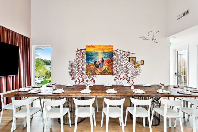 Bright modern coastal dining room with a long rustic wooden communal table and white chairs set with multiple place settings, colorful ocean sunset mural and metal wall art on a white wall, floor-to-ceiling curtains and patio door showing palm trees for a beachy vibe.