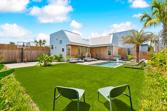 Sunny modern home backyard with pool and patio — outdoor living space featuring palm trees, a basketball hoop, lounge seating and two chairs on a lush green lawn.