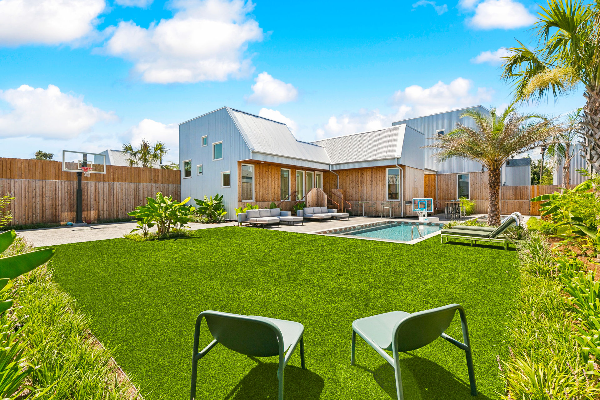 Sunny modern home backyard with pool and patio — outdoor living space featuring palm trees, a basketball hoop, lounge seating and two chairs on a lush green lawn.