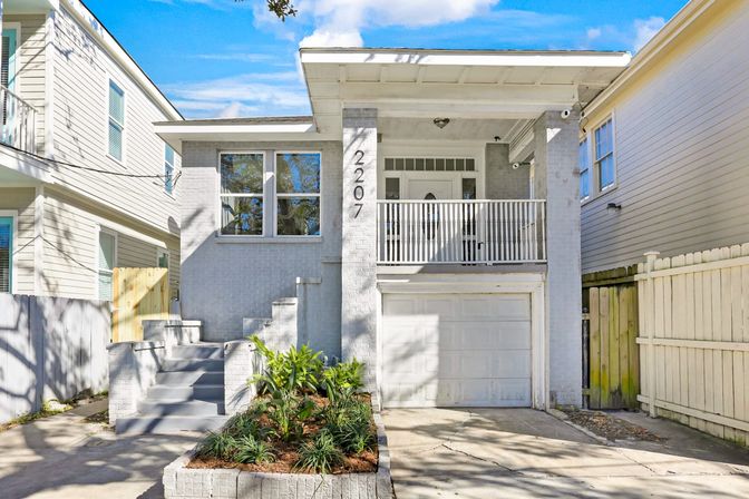 Sunlit gray two-story urban townhouse with raised front porch and balcony, single-car garage, concrete driveway and a small landscaped planter with steps up to the front door.