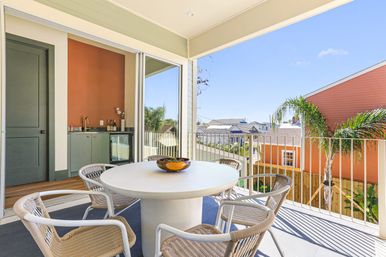 Sunny second-floor balcony dining area with a round white table, four woven chairs and a decorative bowl, sliding glass doors to an indoor wet bar, palm tree and colorful houses in a coastal neighborhood.
