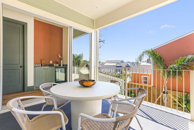 Sunny second-floor balcony dining area with a round white table, four woven chairs and a decorative bowl, sliding glass doors to an indoor wet bar, palm tree and colorful houses in a coastal neighborhood.