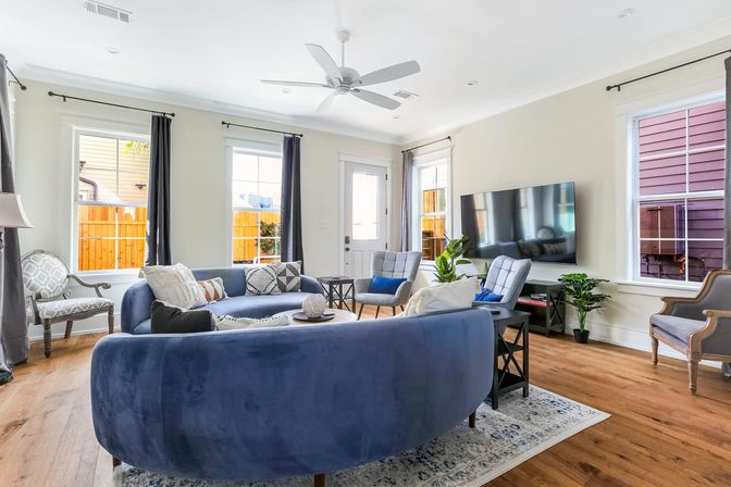 Bright modern living room with curved blue velvet sofa, hardwood floors, large wall-mounted flat-screen TV, multiple windows with dark curtains letting in natural light, ceiling fan, accent chairs, area rug and potted plants.
