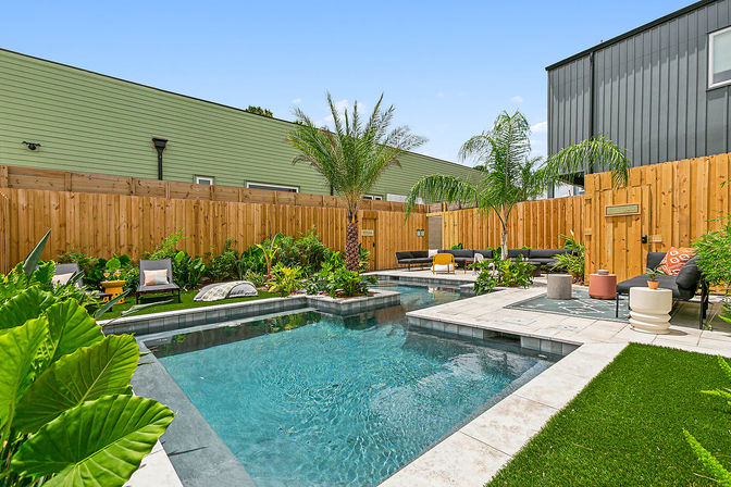 Modern urban backyard with a rectangular turquoise pool, tiled patio, palm trees, wooden privacy fence, lush tropical plants and lounge seating