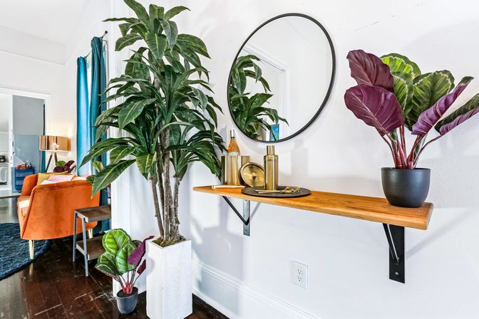 Contemporary apartment entryway with tall potted tropical plant, floating wooden shelf with gold decor and round mirror, orange sofa and teal curtains visible in background.
