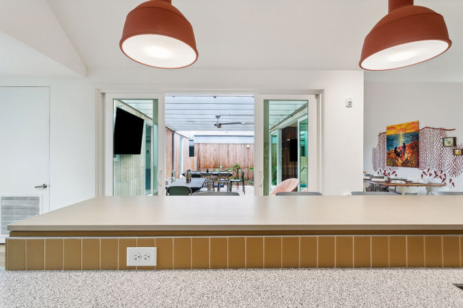 Modern kitchen island with pendant lights overlooking sliding glass doors to a covered outdoor dining patio with wall-mounted TV and colorful wall art