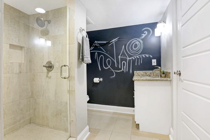 Contemporary home bathroom with glass walk-in shower, beige tile, granite vanity and a black chalkboard accent wall featuring white playful doodle art.