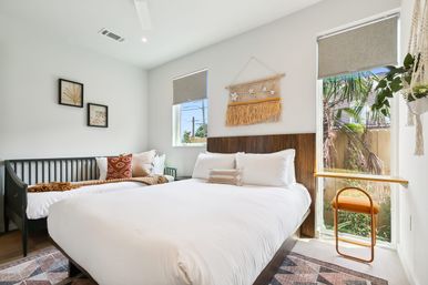 Bright modern boho bedroom with white linens on a queen bed and daybed, wooden slatted headboard, woven wall hanging, large window with tropical plants, small floating desk and orange stool