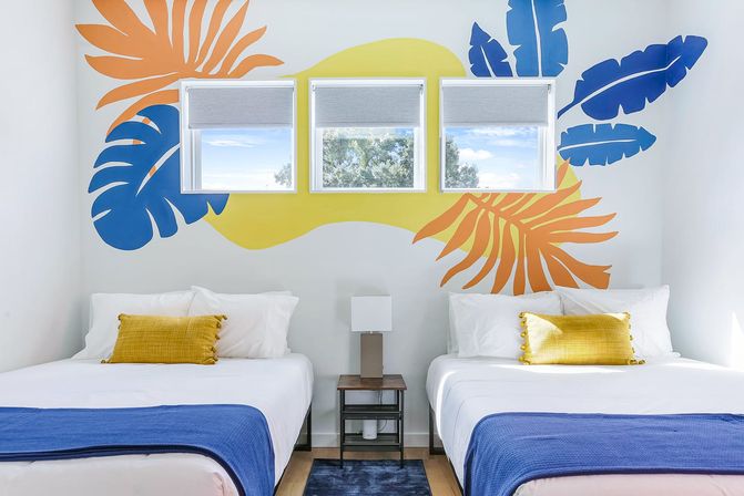 Bright coastal-themed guest room with twin beds, blue and orange tropical leaf mural around three windows, white bedding, blue throws, yellow accent pillows and a central nightstand lamp.