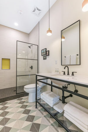 Modern residential bathroom with black-framed glass walk-in shower, matte black fixtures, white vanity on a black metal frame, geometric mint-and-gray floor tiles, pendant lights and neatly folded towels.