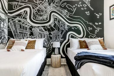 Playful boutique guest room with two beds in white linens, patterned pillows and navy throw, a wooden stump nightstand with lamp and succulent, and a bold black-and-white mural map wall.