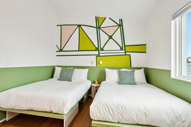 Bright modern twin bedroom with green accent wall and geometric mural, two white beds with patterned pillows, small round nightstand and sunny window