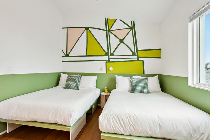 Bright modern twin bedroom with green accent wall and geometric mural, two white beds with patterned pillows, small round nightstand and sunny window