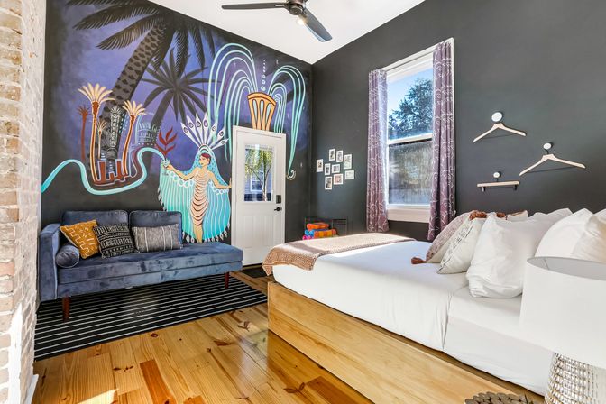 Vibrant eclectic bedroom with a large tropical mural on a dark accent wall, wooden platform bed, velvet navy sofa, striped rug, ceiling fan and sunny window.