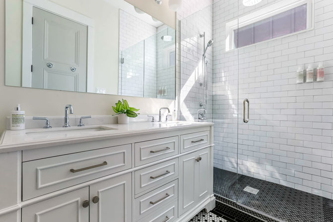 Bright modern white bathroom with double-sink vanity, marble countertop, chrome faucets, glass-enclosed subway-tile shower and black hexagon floor tile