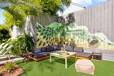 Sunlit coastal backyard lounge with gray corner sofa, mint-green coffee table on artificial turf, ocean-themed fence mural of fish and crab, palm plants, pouf and a wine and charcuterie spread.
