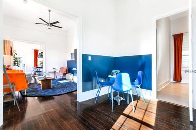 Sunlit open-plan living room and breakfast nook with a blue half-wall, round table and bright blue chairs, orange sofa, navy rug, ceiling fan and dark hardwood floors.