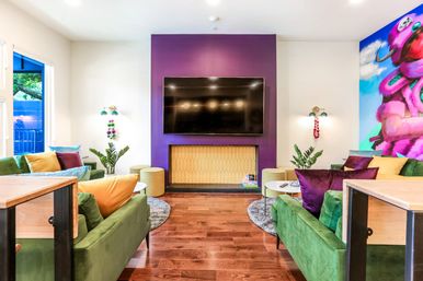 Vibrant modern living room with purple TV wall and large flat-screen, green velvet sofas with colorful pillows, hardwood floors, round rugs, plants and a playful pink elephant mural.