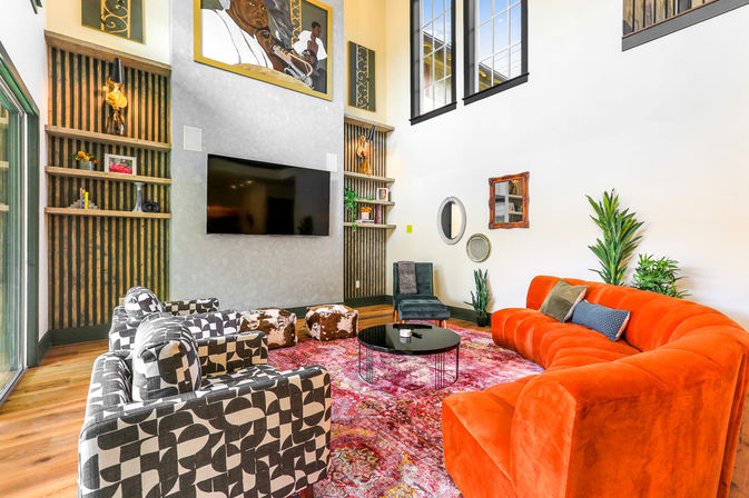 Bright modern living room with double-height ceiling, vibrant orange velvet curved sofa, black-and-white geometric armchairs, wall-mounted TV, pink patterned rug, cowhide poufs, tall windows and indoor plants.