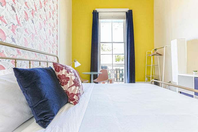 Sunny city apartment bedroom with a yellow accent wall and pink bird wallpaper, white bed styled with navy and floral pillows, small round table and pink chair by a tall window opening to a balcony with decorative wrought-iron railing.
