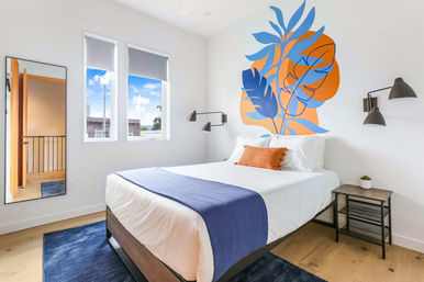 Bright modern bedroom with white walls and hardwood floor, queen bed dressed in white linens with a navy throw and orange accent pillow, bold blue-and-orange botanical wall mural behind the headboard, black wall sconces, small wooden nightstand with plant, full-length mirror and two windows with natural light.