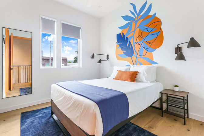 Bright modern bedroom with white walls and hardwood floor, queen bed dressed in white linens with a navy throw and orange accent pillow, bold blue-and-orange botanical wall mural behind the headboard, black wall sconces, small wooden nightstand with plant, full-length mirror and two windows with natural light.