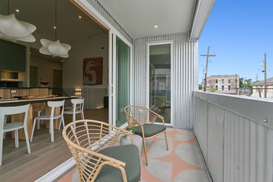 Urban apartment balcony with two rattan lounge chairs and small table on geometric tile, sliding glass door to open-plan dining area with white chairs and sculptural pendant lights, corrugated metal siding and distant city buildings