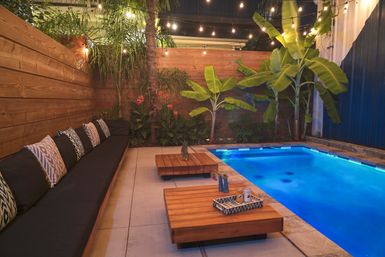 Cozy modern backyard patio with glowing blue rectangular pool, long cushioned bench with patterned pillows, low wooden coffee tables, tropical banana plants and hanging string lights at dusk.