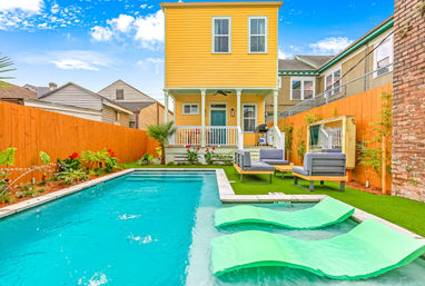 Sunny backyard with a bright yellow two-story house and porch overlooking a rectangular turquoise swimming pool, mint green lounger pads on the shallow ledge, modern gray patio sofas on artificial turf, orange wooden fence and tropical plants under a blue sky.