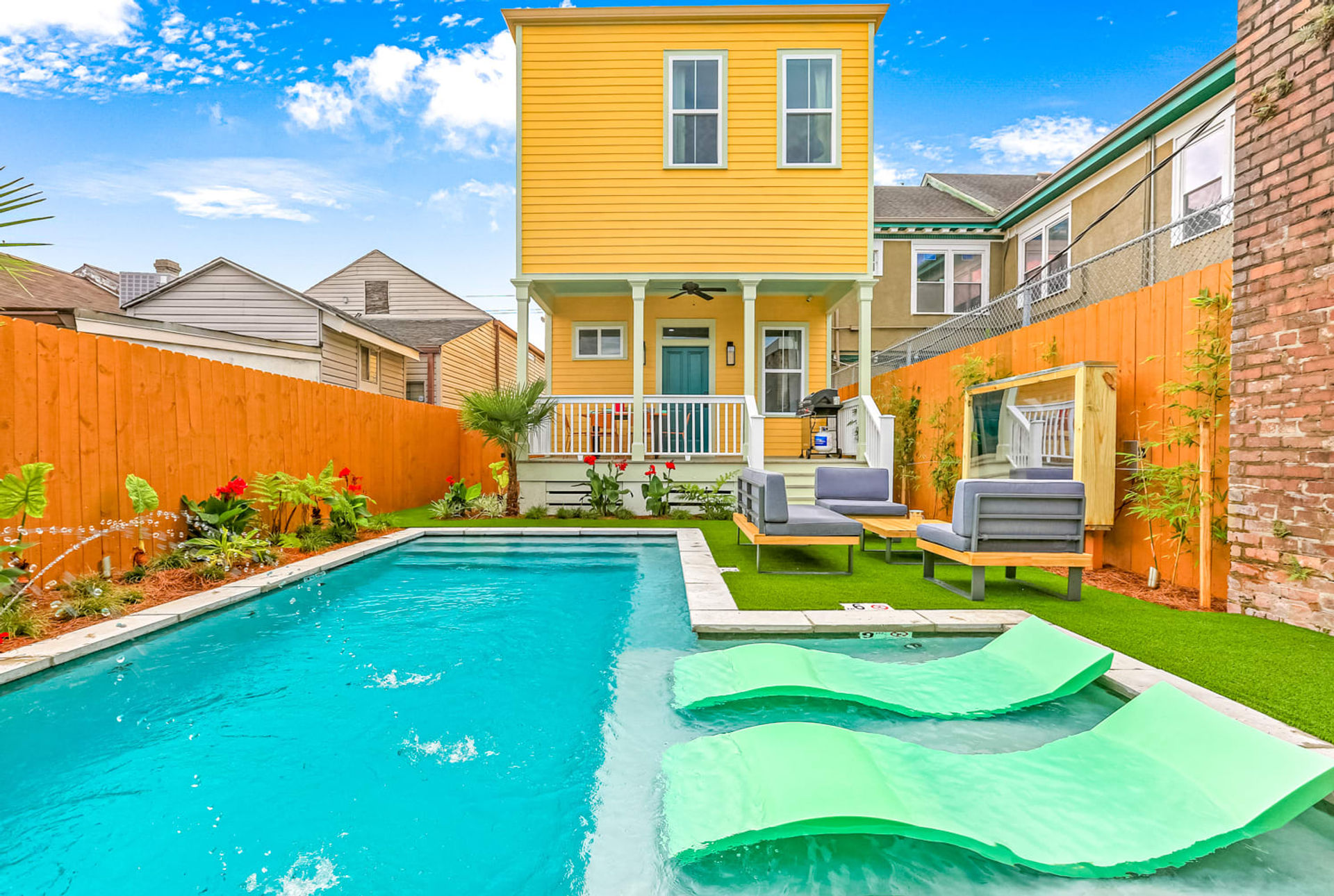 Sunny backyard with a bright yellow two-story house and porch overlooking a rectangular turquoise swimming pool, mint green lounger pads on the shallow ledge, modern gray patio sofas on artificial turf, orange wooden fence and tropical plants under a blue sky.