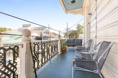 Sunny second-story balcony with ornate wrought-iron railing, blue painted floor, wicker chairs, cushioned sofa, potted plants and decorative eaves overlooking a small commercial street.