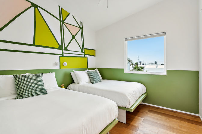 Cheerful modern twin bedroom with crisp white bedding, green geometric accent wall and throw pillows, hardwood floor, and a window framing palm trees and a distant city skyline under a blue sky.