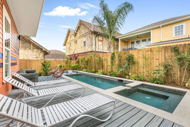 Sunny urban backyard with narrow rectangular pool and built-in spa, grey wood deck lined with white lounge chairs, tropical plants and wooden privacy fence beside neighboring houses