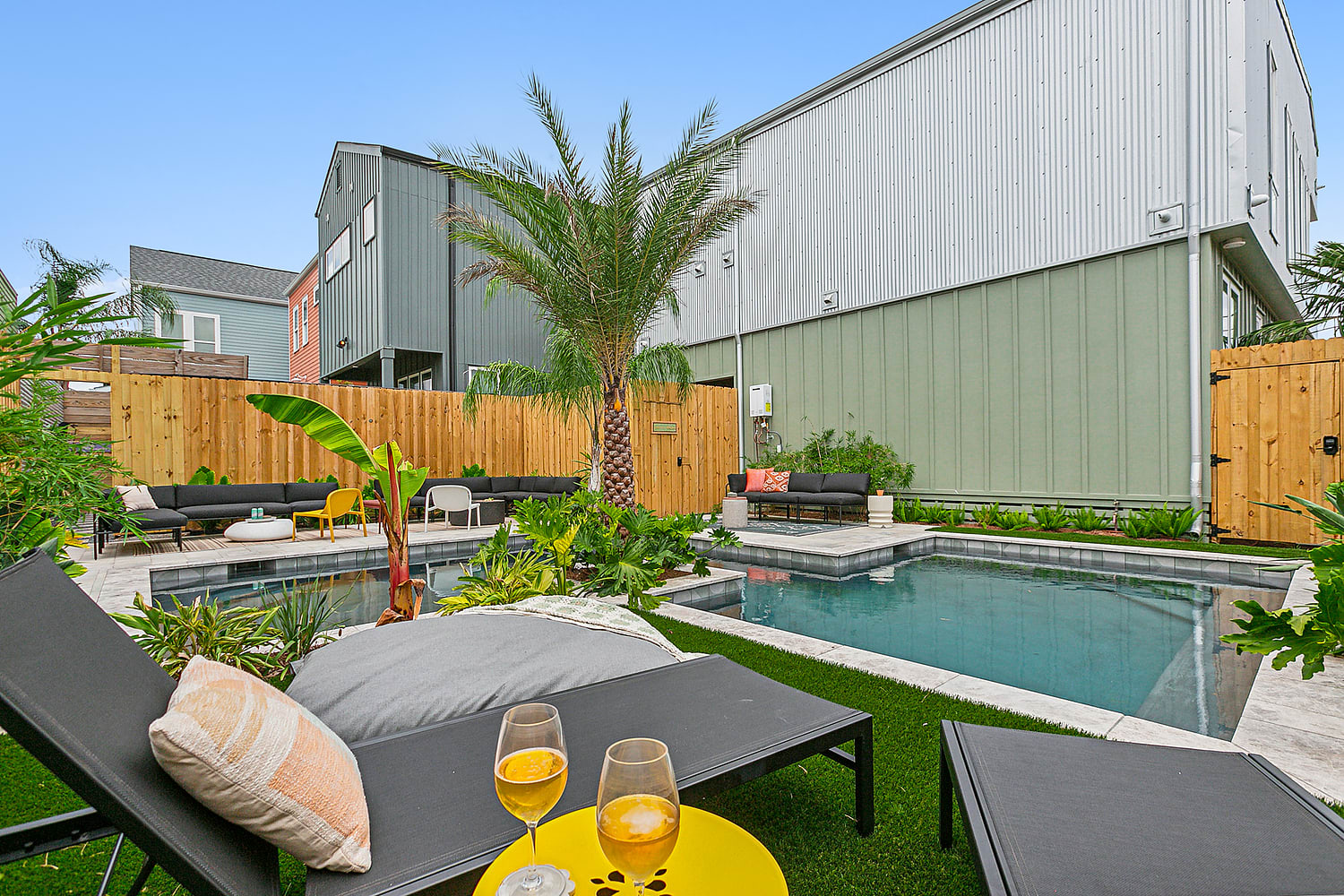 Modern private backyard pool and patio with a central palm tree, tropical landscaping, black lounge chairs, a yellow side table with two drinks, sectional seating and a wooden fence next to contemporary townhomes.