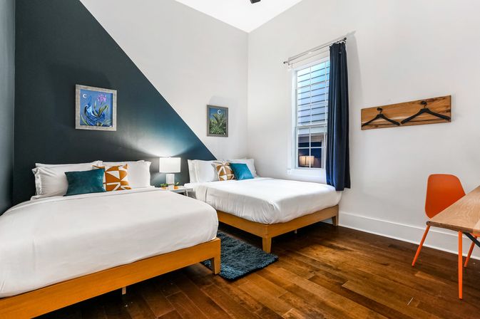 Bright modern guest room with two wooden-framed double beds, white bedding and colorful throw pillows, navy diagonal accent wall with framed art, bedside lamp, hardwood floors, window with navy curtain, orange desk chair and wall-mounted wooden coat rack.