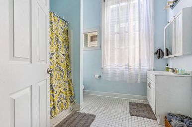 Cheerful light-blue bathroom with a yellow patterned shower curtain, sheer window curtains, white vanity, and tiled floor.