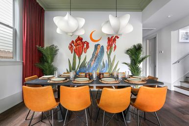Bright modern dining room with eight orange leather chairs around a black table set with woven placemats and glassware; a colorful floral wall mural with red irises, blue abstract shapes and a playful orange crescent moon; white sculptural pendant lights, potted palms, dark wood floors and red drapery.