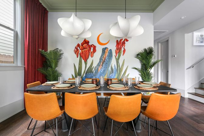 Bright modern dining room with eight orange leather chairs around a black table set with woven placemats and glassware; a colorful floral wall mural with red irises, blue abstract shapes and a playful orange crescent moon; white sculptural pendant lights, potted palms, dark wood floors and red drapery.